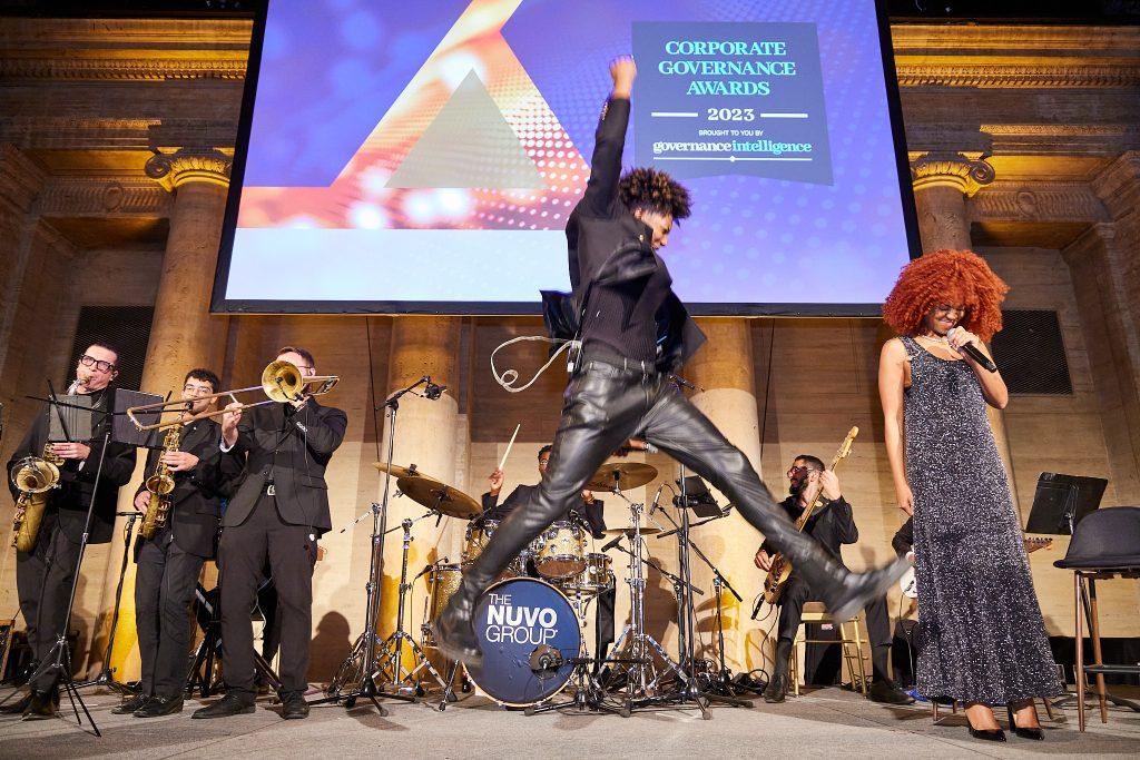 Lead performer mid-jump with vocalist and horn section at a corporate awards show
