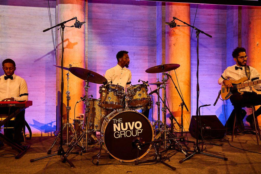 Keyboard, drums, and bass trio performing under warm stage lighting